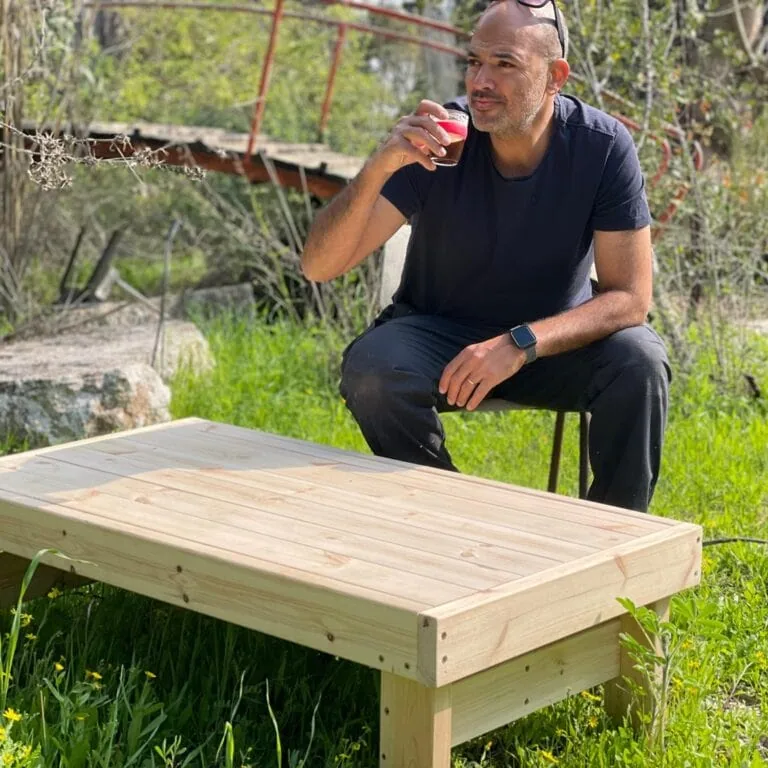 שולחן קפה לגינה מעץ דגם "דולב" בחוץ על דשא, אדם יושב ברקע
