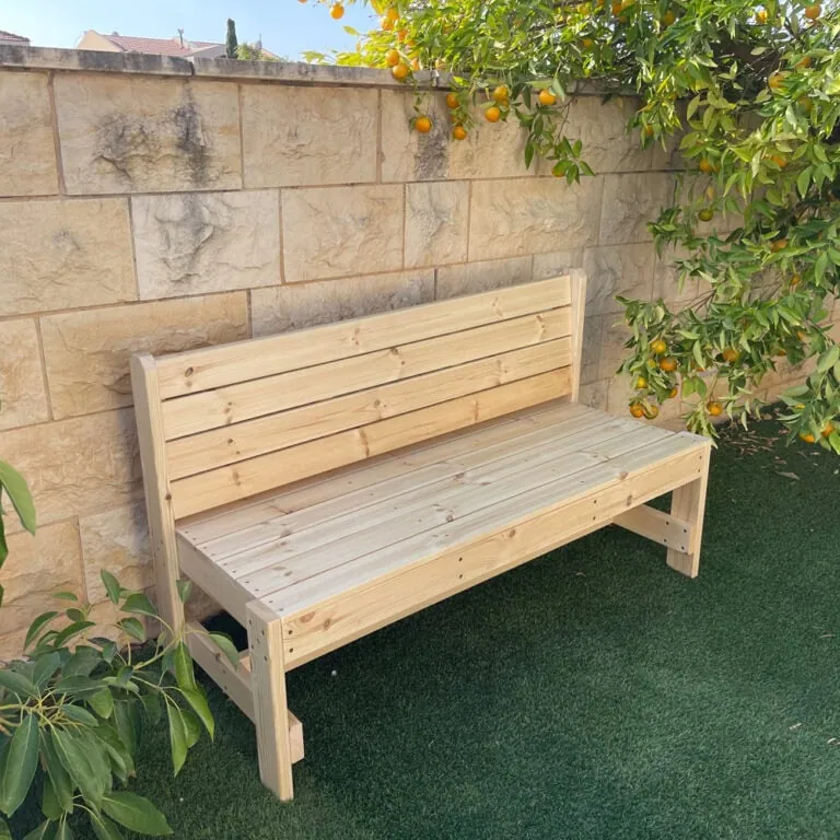 ספסל עץ עם משענת דגם בייסיק ניצב מול קיר אבן וגינה