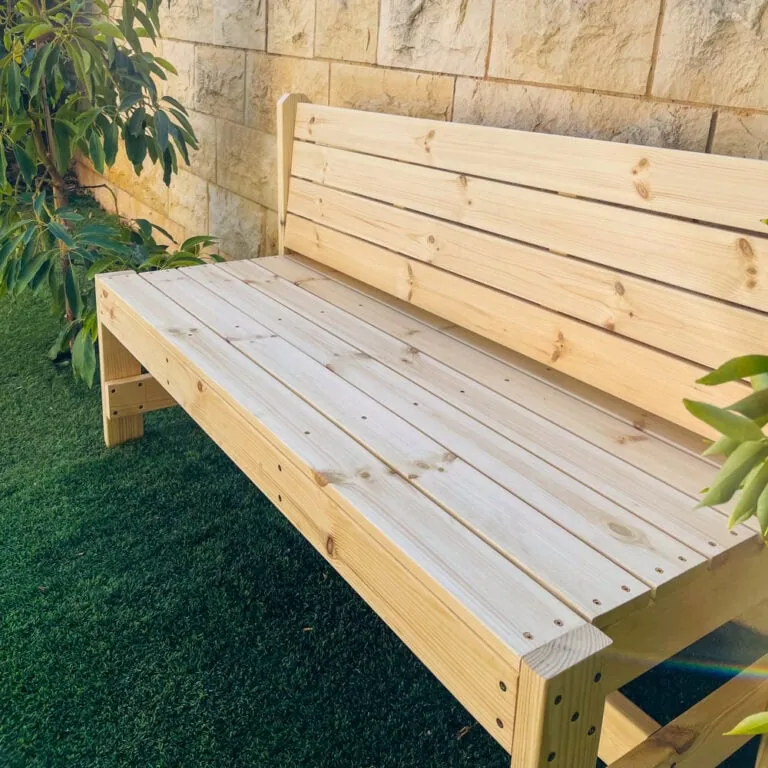 ספסל עץ אורן עם משענת דגם "בייסיק" על דשא מול קיר אבן