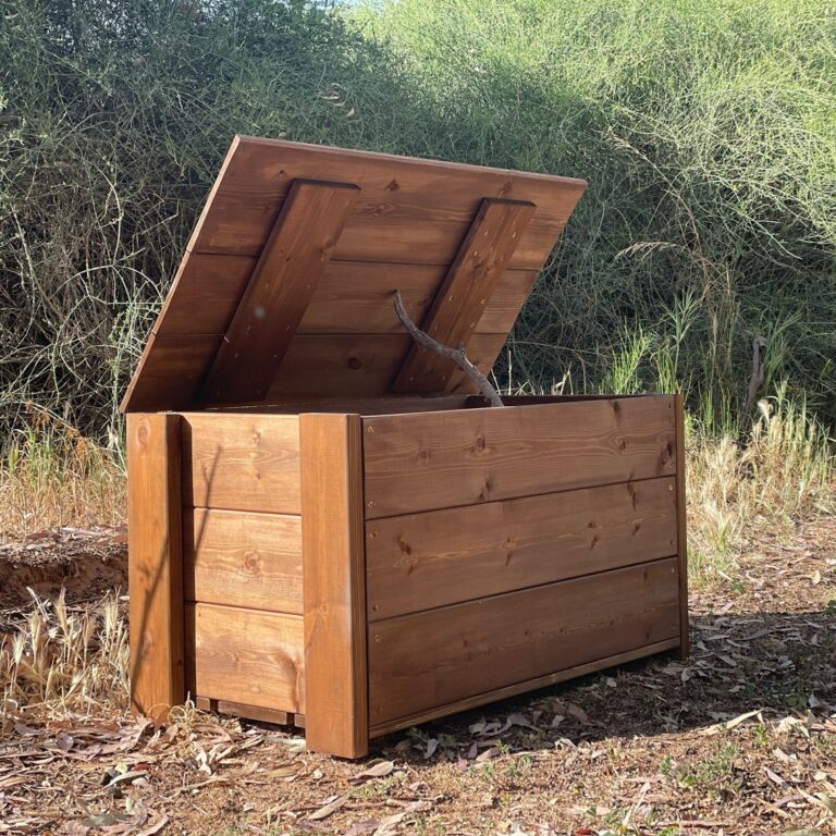 ספסל אחסון מעץ לגינה ולמרפסת עם מכסה פתוח בגינה על רקע שיחים